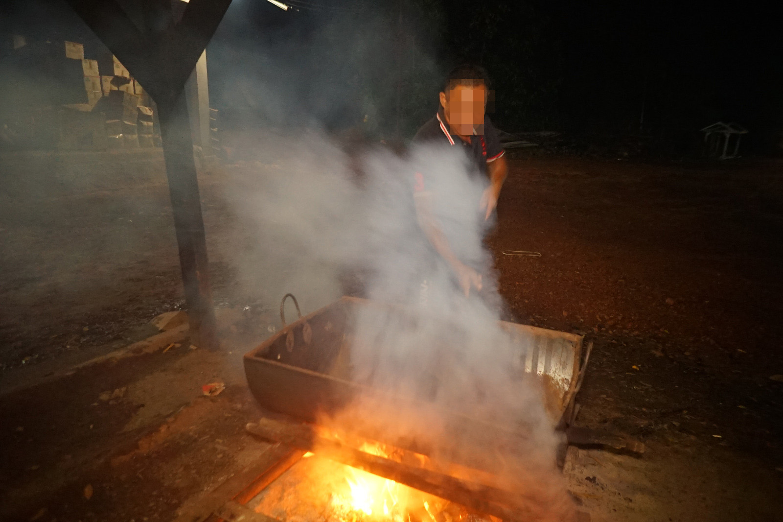 A man with a blurred face stands over an oil drum cut in half and placed over a fire